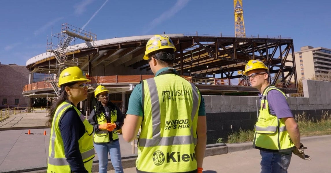 Four construction professionals wearing PPE stand in a circle and talk with a construction project in the background. Four construction professionals wearing PPE stand in a circle and talk with a construction project in the background.