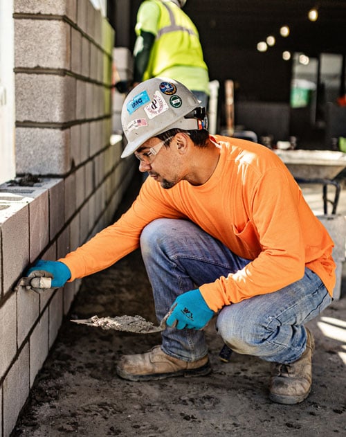 A mason holding a trowel squats next to an examines the concrete block wall he is building.