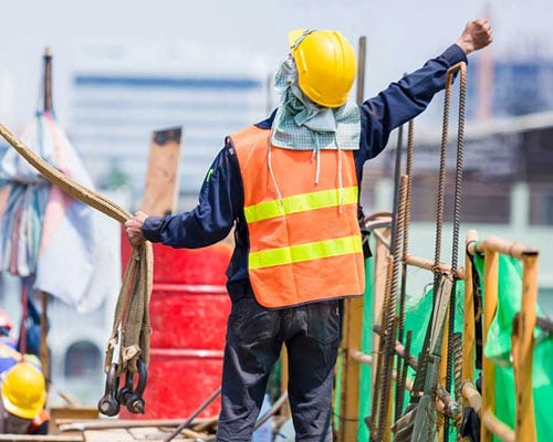 A signal person holds cables in one hand and has the other hand outstretched in a fist, sending a hand signal to a crane operator.