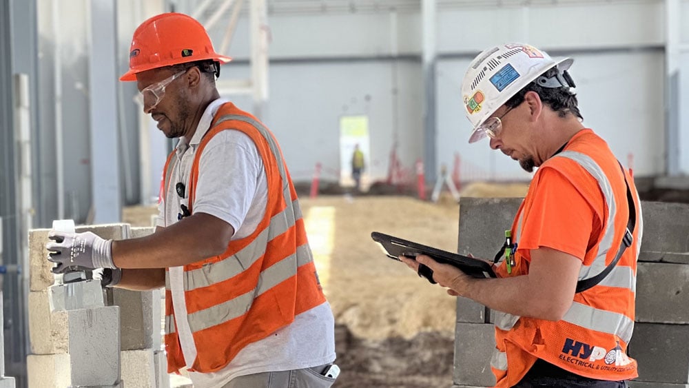 One construction professional places brick while a supervisor observes and makes note on a tablet. One construction professional places brick while a supervisor observes and makes note on a tablet.