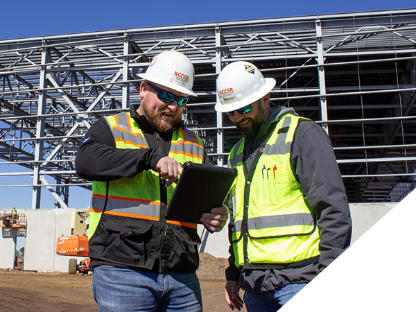A construction supervisor and another worker look at a tablet in front of a construction project. A construction supervisor and another worker look at a tablet in front of a construction project.